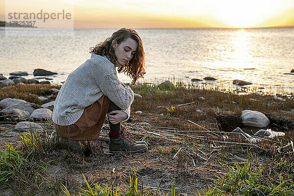 Junge Frau sitzt bei Sonnenuntergang auf einem Felsen vor dem Meer