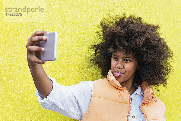 Mädchen streckt die Zunge heraus und macht ein Selfie mit dem Smartphone vor gelbem Hintergrund