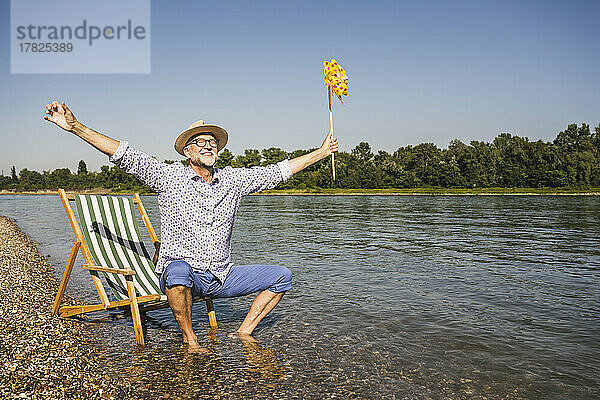 Lächelnder Mann mit ausgestreckten Armen hält ein Windradspielzeug aus Papier am Flussufer