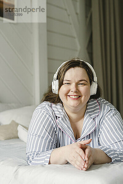 Lächelnde Frau  die auf dem Bett liegt und Musik über drahtlose Kopfhörer hört