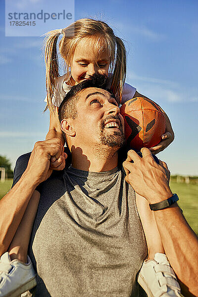 Glücklicher Mann blickt Tochter an  die mit einem Rugbyball auf den Schultern sitzt