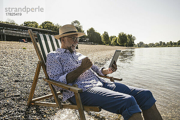 Glücklicher Mann mit Tablet-PC sitzt auf Liegestuhl am Flussufer