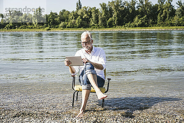 Smiling man using tablet PC sitting on chair at beach