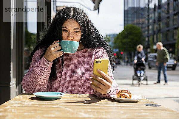 Junge Frau trinkt Kaffee mit Smartphone und sitzt im Straßencafé