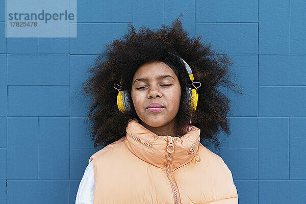 Mädchen mit geschlossenen Augen hört Musik über kabellose Kopfhörer vor einer blauen Wand