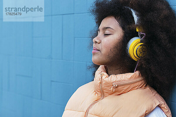 Mädchen mit geschlossenen Augen hört Musik und lehnt an einer blauen Wand