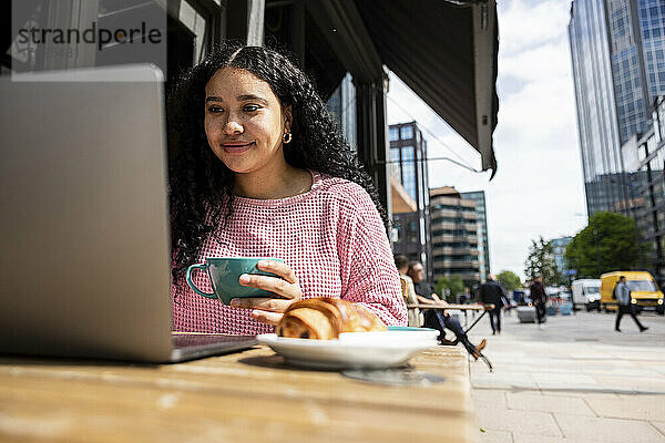 Lächelnde Frau mit Kaffeetasse und Laptop im Straßencafé