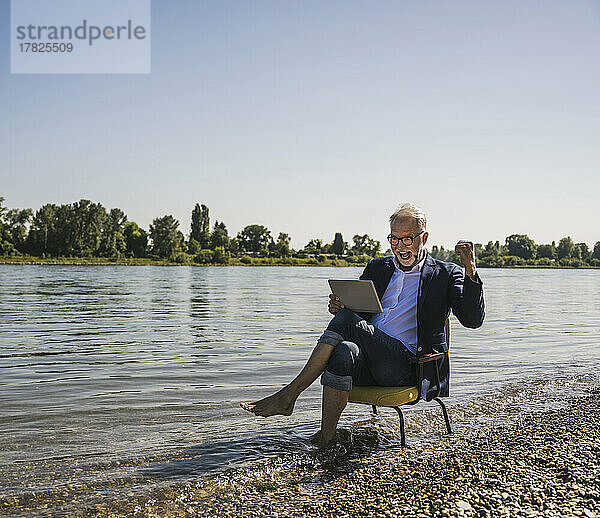 Glücklicher Mann mit Tablet-PC sitzt auf einem Stuhl am Flussufer