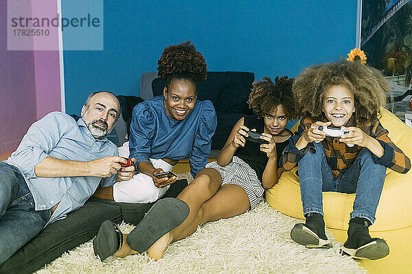 Lächelnde Frau mit Mobiltelefon sitzt neben Mann und Kindern und spielt zu Hause Videospiele
