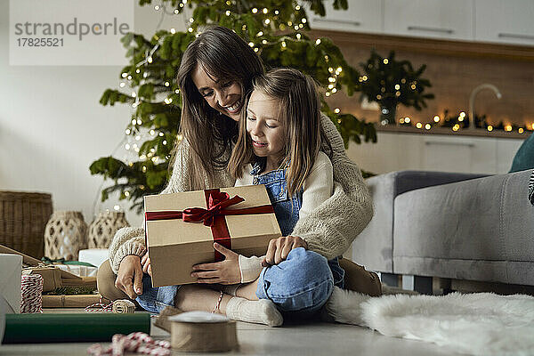 Mädchen hält Weihnachtsgeschenk und sitzt mit Mutter zu Hause auf dem Boden