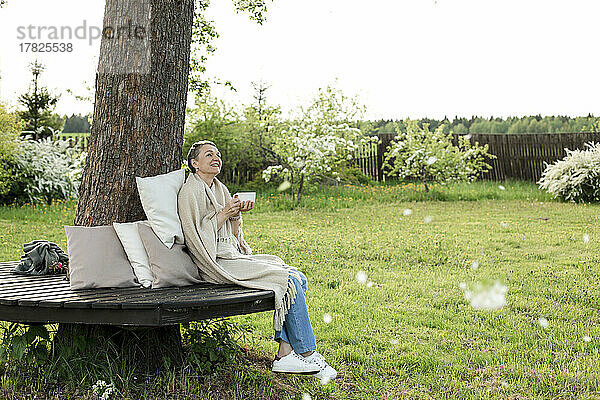 Lächelnde Frau  in Decke gehüllt  mit Tasse auf Bank um Baumstamm sitzend