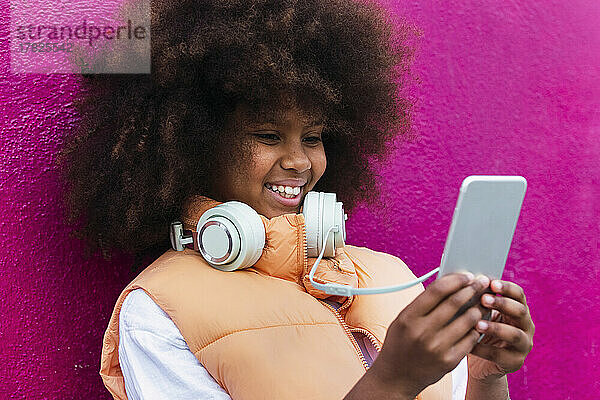 Mädchen mit Kopfhörern und Mobiltelefon an rosa Wand gelehnt