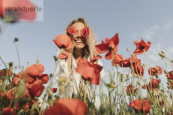 Glückliche Frau bedeckt Augen mit Blumen im Mohnfeld