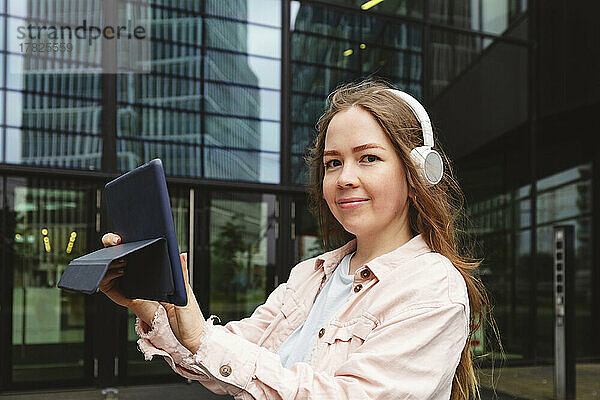 Glückliche Frau mit kabellosen Kopfhörern steht mit Tablet-PC vor dem Gebäude