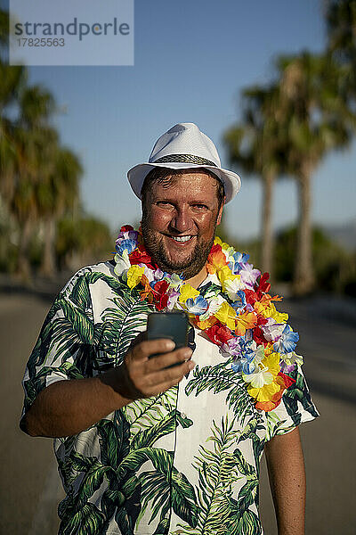Glücklicher Mann mit Blumengirlande und Smartphone auf der Straße