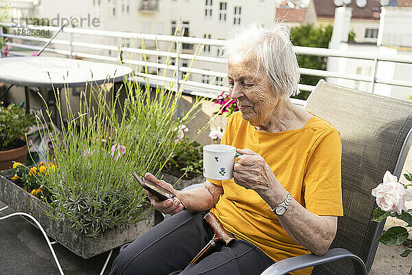 Lächelnde ältere Frau hält Kaffeetasse in der Hand und blickt auf ihr Mobiltelefon auf dem Balkon