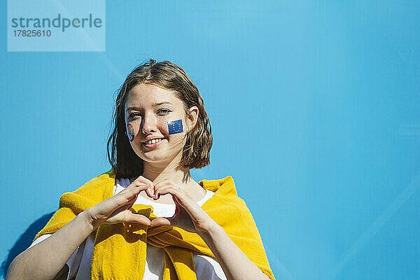 Lächelndes Teenager-Mädchen mit Friedenssymbol und EU-Farbe auf den Wangen gestikuliert in Herzform vor einer blauen Wand