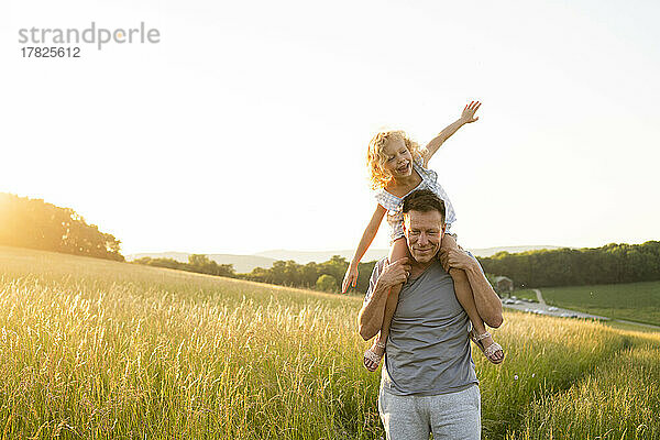 Lächelnder Mann mit Tochter sitzt mit ausgestreckten Armen auf dem Feld