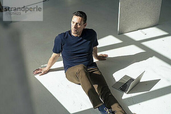 Geschäftsmann mit Laptop sitzt auf dem Boden im Sonnenlicht im Büro