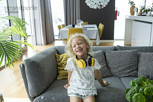 Fröhliches Mädchen mit kabellosen Kopfhörern sitzt zu Hause auf dem Sofa