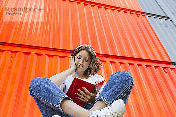 Teenagermädchen mit rotem Notizbuch sitzt vor orangefarbenem Frachtcontainer