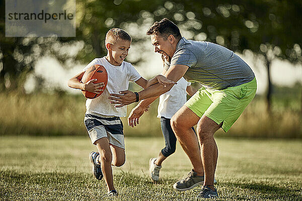 Fröhlicher Mann jagt Sohn  der mit Rugbyball auf dem Sportplatz rennt