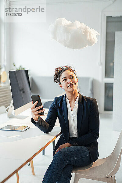 Lächelnde junge Geschäftsfrau mit Mobiltelefon und Blick auf die schwebende Wolke im Büro