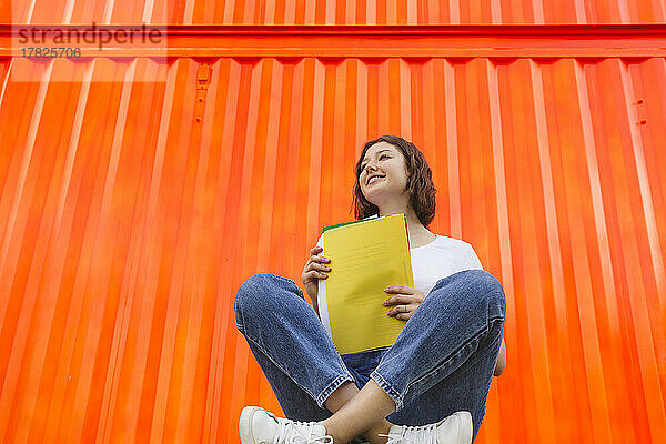 Smiling teenage girl with yellow file folder sitting cross-legged in front of orange cargo container