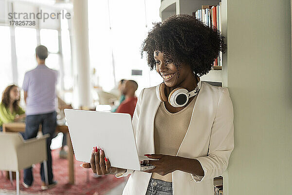 Lächelnde berufstätige Frau benutzt Laptop und stützt sich im Büro auf ein Regal