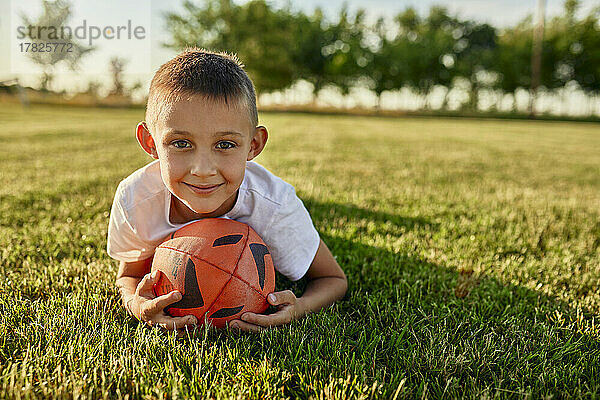 Lächelnder Junge mit Rugbyball  der an einem sonnigen Tag auf dem Sportplatz liegt