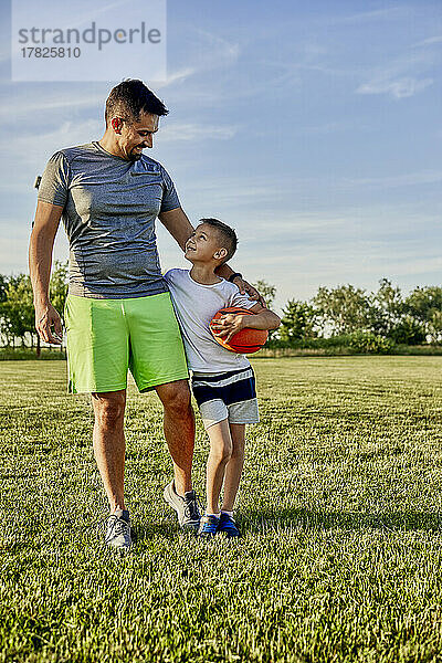 Lächelnder Mann geht mit Sohn und hält Rugbyball auf dem Sportplatz