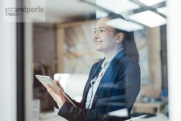 Lächelnde Geschäftsfrau mit Tablet-PC  die durch das Fenster schaut