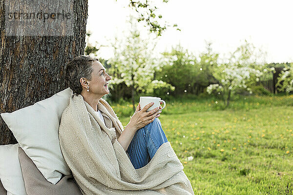 Glückliche Frau  in eine Decke gehüllt und mit einer Tasse sitzend  auf einer Bank um einen Baumstamm sitzend