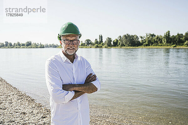 Lächelnder leitender Ingenieur mit grünem Schutzhelm steht mit verschränkten Armen am Flussufer