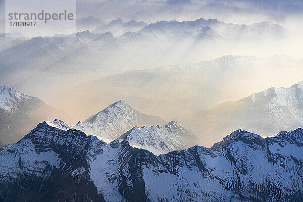 Sonnenstrahlen durch Nebel über den schneebedeckten Gipfeln der Lepontiner und Tessiner Alpen bei Sonnenuntergang Blick aus dem Flugzeug im Flug  Schweiz  Europa