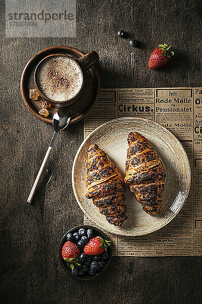 Croissants auf Teller dazu eine Tasse Kaffee und frische Beeren auf Holztisch