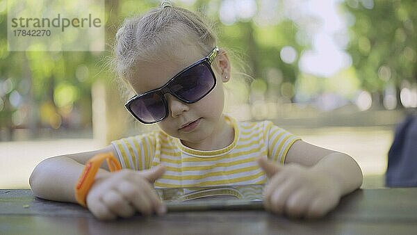 Kleines Mädchen mit Sonnenbrille lernt Fremdsprache durch Wiederholung von Wörtern aus dem Handy. Close-up-Porträt von Kind Mädchen sitzt in Stadtpark und lernen fremde Sprache mit Handy. Odessa  Ukraine  Europa