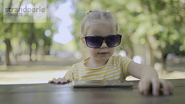 Kleines Mädchen mit Sonnenbrille lernt Fremdsprache durch Wiederholung von Wörtern aus dem Handy. Close-up-Porträt von Kind Mädchen sitzt in Stadtpark und lernen fremde Sprache mit Handy. Odessa  Ukraine  Europa
