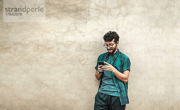 Happy gut aussehend Kerl lehnt sich gegen die Wand mit seinem Handy  glücklich latin Mann mit seinem Handy lehnt sich gegen eine Wand mit Kopie Raum  Person lächelnd Telefon mit Platz für Text