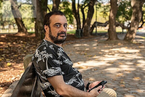 Ein Mann sitzt auf einer Bank im Park mit seinem Handy in der Hand und schaut in die Kamera