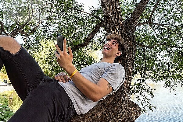 Junger Mann lehnt an einem Baumstamm  lacht und benutzt sein Smartphone