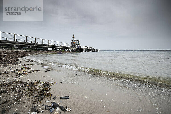 Bick vom Strand auf die Seebrücke von Glücksburg mit dem DLRG-Rettungsturm unter grauem Regenhimmel. Die Tropfen malen Muster auf die Wasseroberfläche der Flensburger Förde