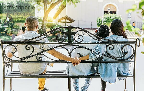 Untreue Frau sitzt händchenhaltend mit einem anderen Mann  während ihr Freund sie umarmt. Untreues Paar hält Hände auf einer Parkbank. Konzept des Liebesdreiecks und der Untreue