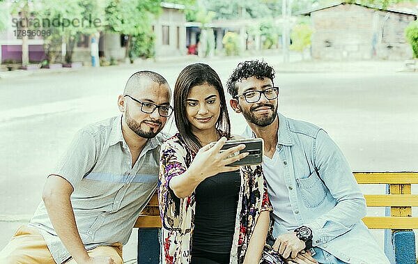 Drei Freunde machen ein Selfie auf einer Parkbank  Drei lächelnde Freunde sitzen auf einer Bank und machen ein Selfie. Vorderansicht von drei glücklichen Freunden  die auf einer Bank sitzend ein Selfie machen
