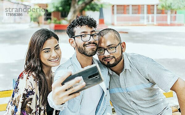 Drei lächelnde Freunde sitzen auf einer Bank und machen ein Selfie. Vorderansicht von drei glücklichen Freunden  die auf einer Bank sitzend ein Selfie machen. Drei Freunde machen ein Selfie auf einer Parkbank