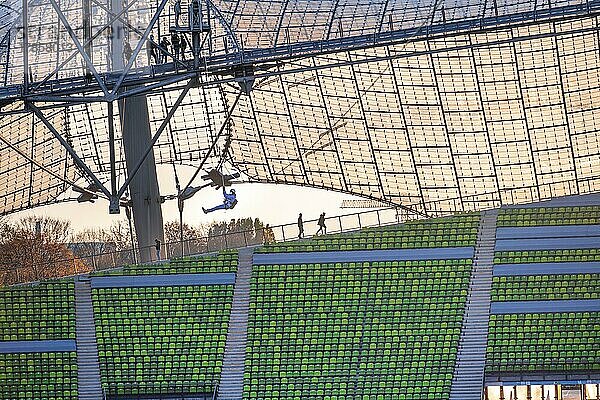 Frau schwingt an einer Zip-Line über dem Olympiastadion  Olympiastadion mit Zeltdach bei Sonnenuntergang  Olympiapark  München  Bayern  Deutschland  Europa