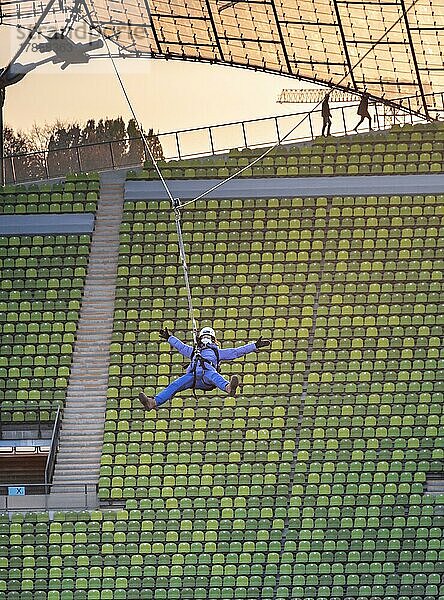 Frau schwingt an einer Zip-Line über dem Olympiastadion  Olympiastadion mit Zeltdach bei Sonnenuntergang  Olympiapark  München  Bayern  Deutschland  Europa