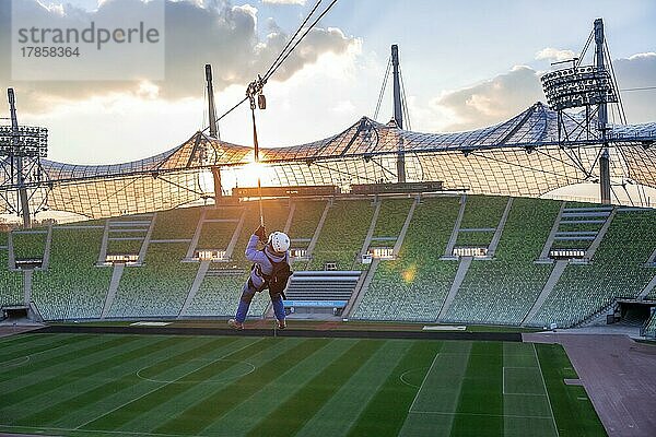 Frau schwingt an einer Zip-Line über dem Olympiastadion  Olympiastadion mit Zeltdach bei Sonnenuntergang  Olympiapark  München  Bayern  Deutschland  Europa