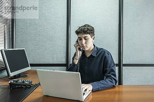Junger Mann arbeitet an seinem Laptop  während er im Büro telefoniert