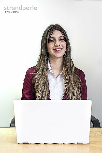 Schöne lächelnde Frau arbeitet an ihrem weißen Laptop und schaut in die Kamera